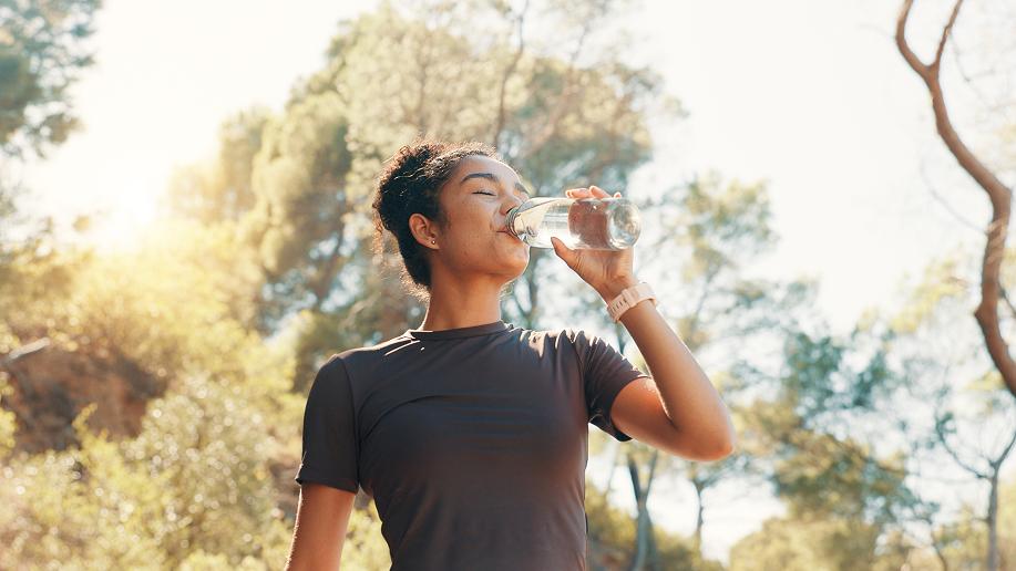 Woman in black t-shirt drinking water from a bottle outdoors among trees on a sunny day.